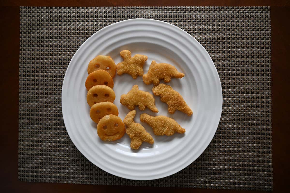 Top down image of dino chicken nuggets cooked and served on a white plate beside smiles fries.