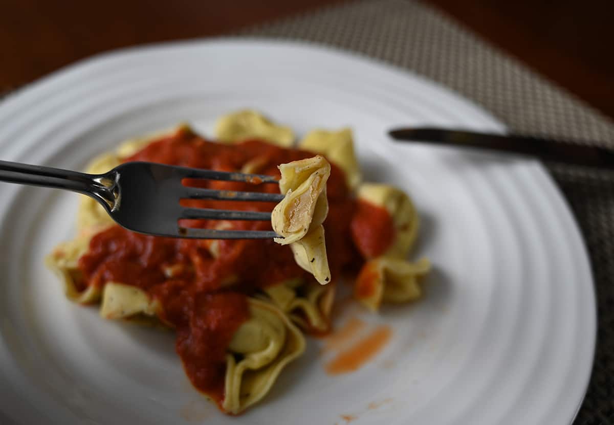 Image of a fork with a tortelloni close to the camera cut in half so you can see the filling. In the background is a plate of tortelloni with red sauce on it.