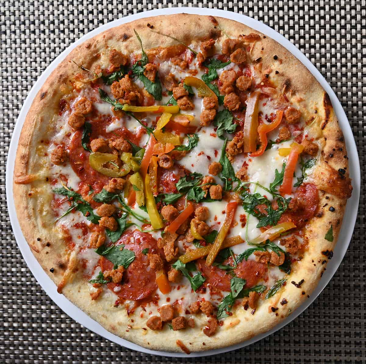 Top down image of a sausage, salami, arugula and grilled peppers pizza baked and served whole on a white plate.
