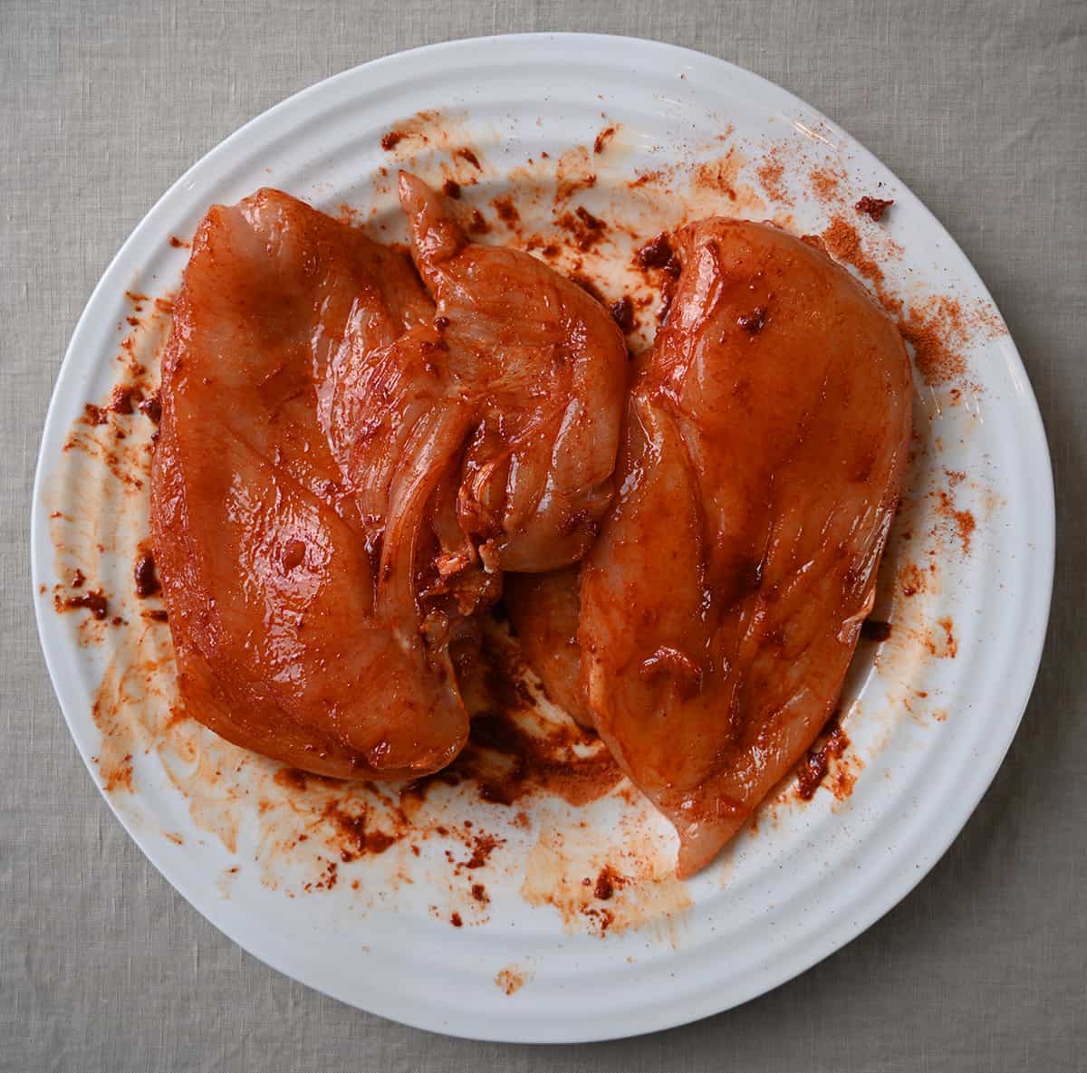 Image of two raw chicken breasts with sriracha seasoning rubber on them, sitting on a plate.
