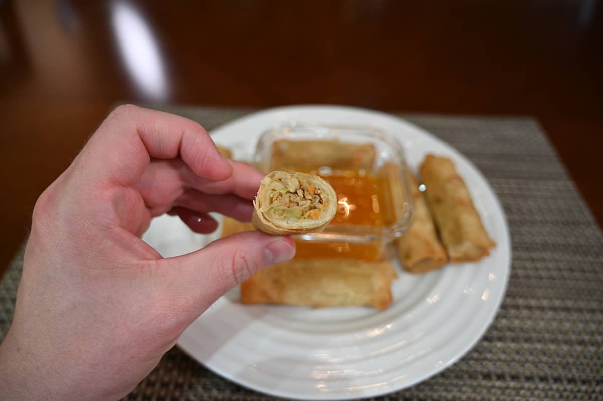 Image of a hand holding one crispy spring roll with a bite taken out of it so you can see the center, in the background is a plate of spring rolls and a bowl of plum sauce.