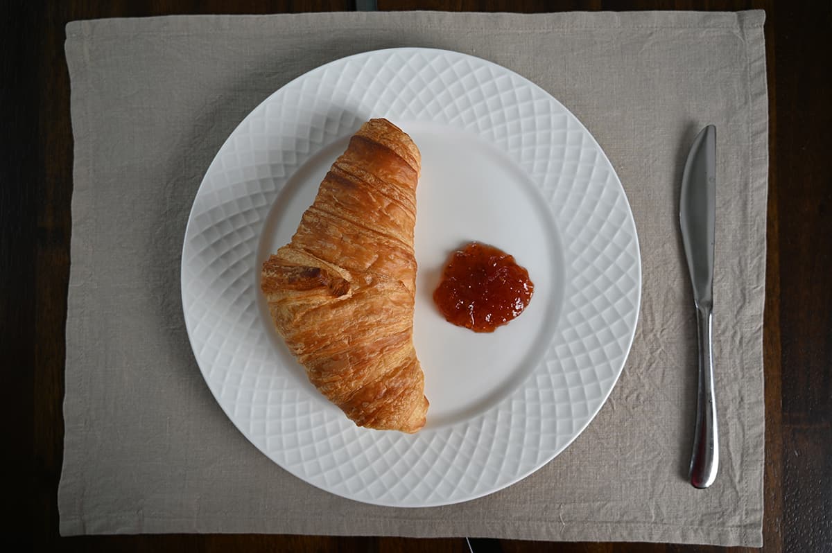 Top down image of a butter croissant served on a white plate with a dollop of jam beside it. 