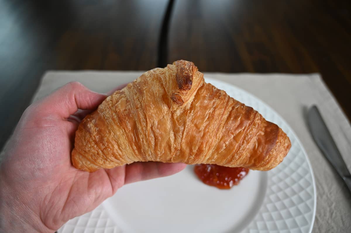 Closeup image of a hand holding one full croissant close to the camera with a white plate in the background.
