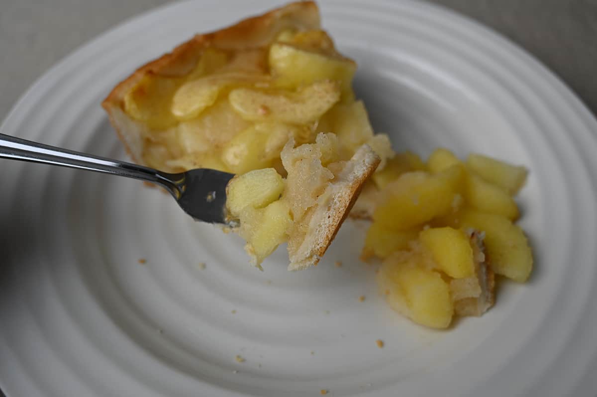 Closeup image of a fork with a bit of cake and apple filling on the fork. In the background is a piece of apple cake on a plate. 