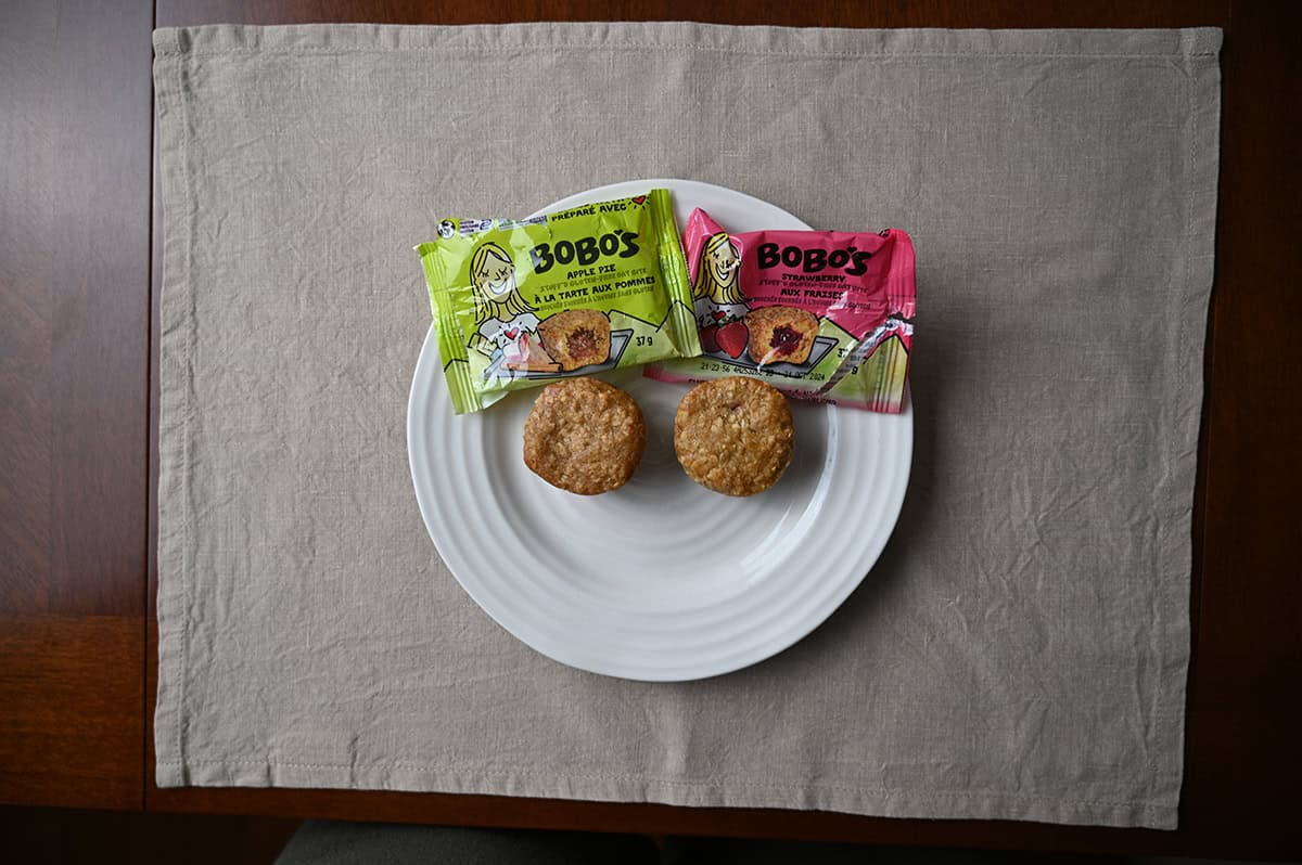 Top down image of two oat bites unpackaged served on a white plate sitting beside the wrappers for the apple and strawberry oat bites.
