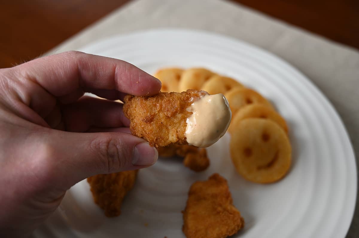 Closeup image of a hand holding a chicken nugget dipped in sauce close to the camera with a plate of fries in the background.
