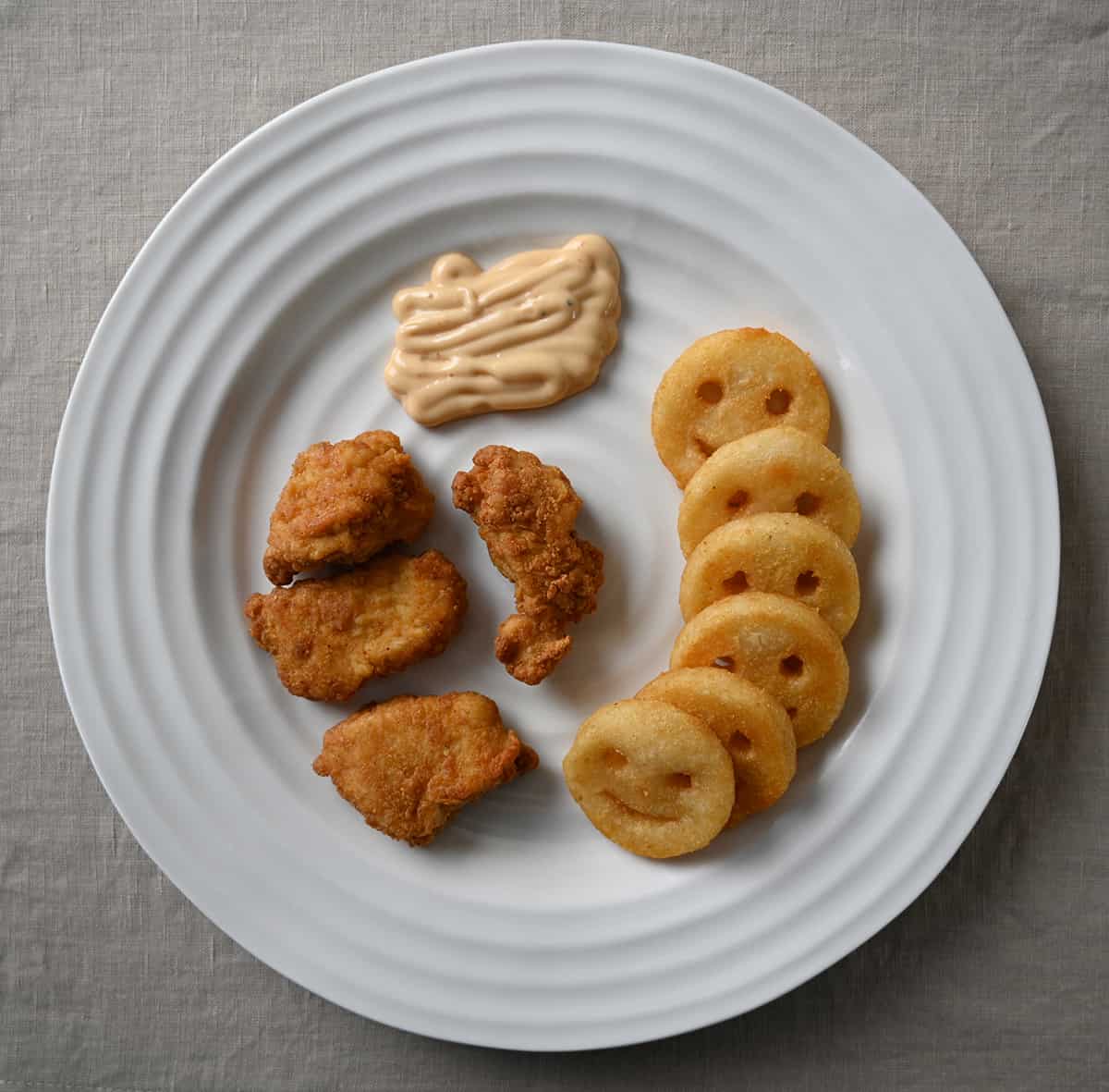 Top down image of a plate with chicken breast chunks on it beside smiley fries and dip. 