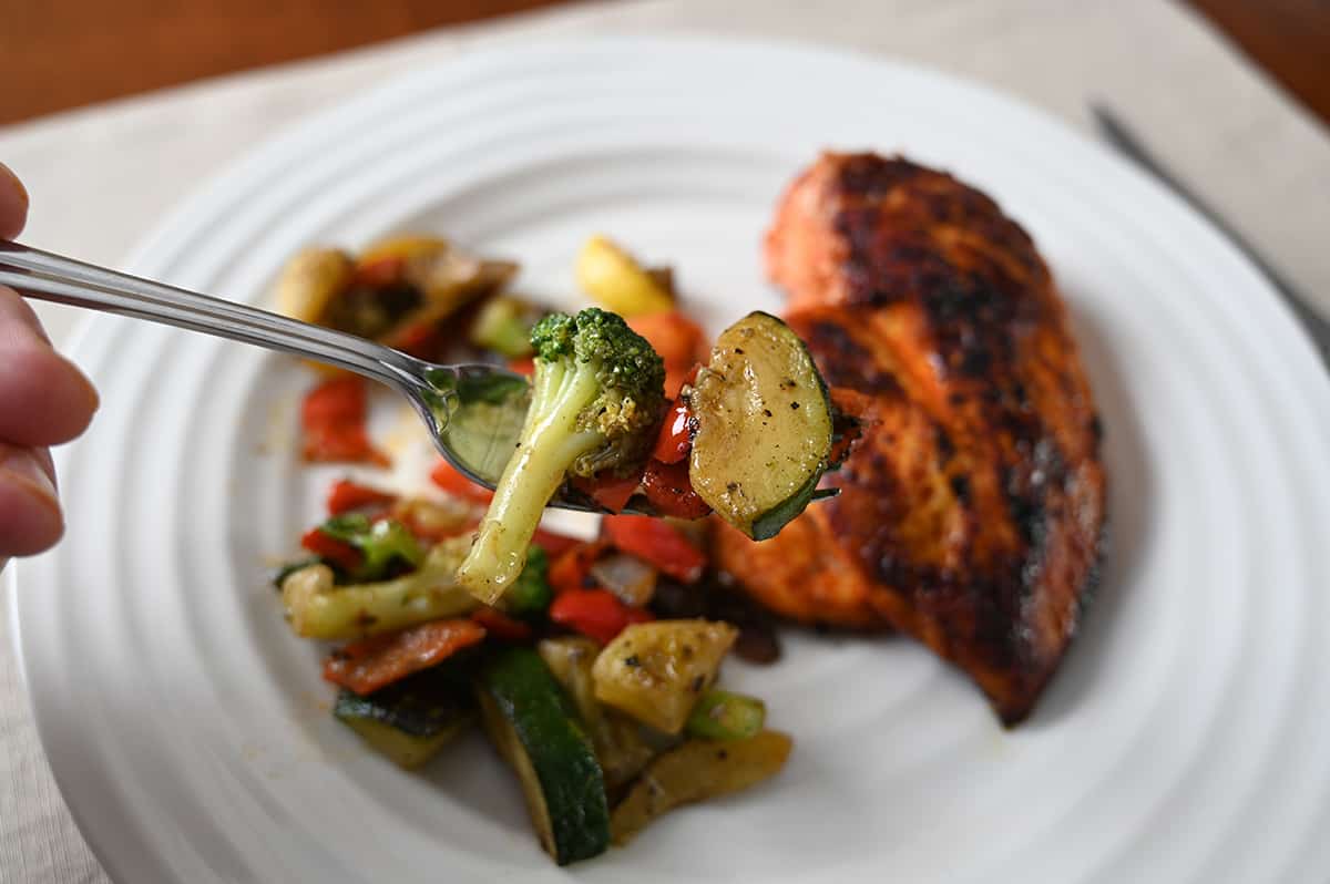 Closeup image of a fork with a piece of broccoli and zucchini on the fork and a plate of chicken and roasted vegetables in the background.