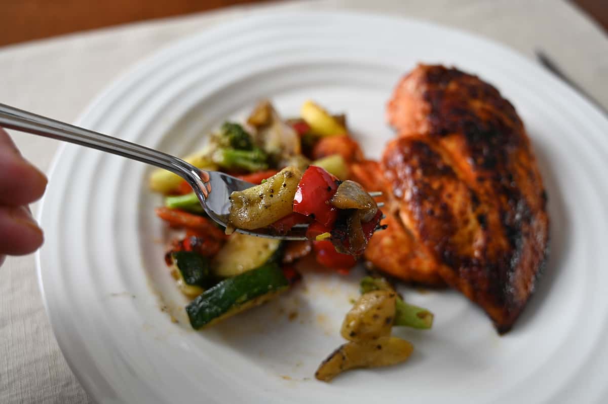 Closeup image of a fork with with red pepper, zuchinni and red onion on it. In the background of the image is a plate with vegetables beside a chicken breast.