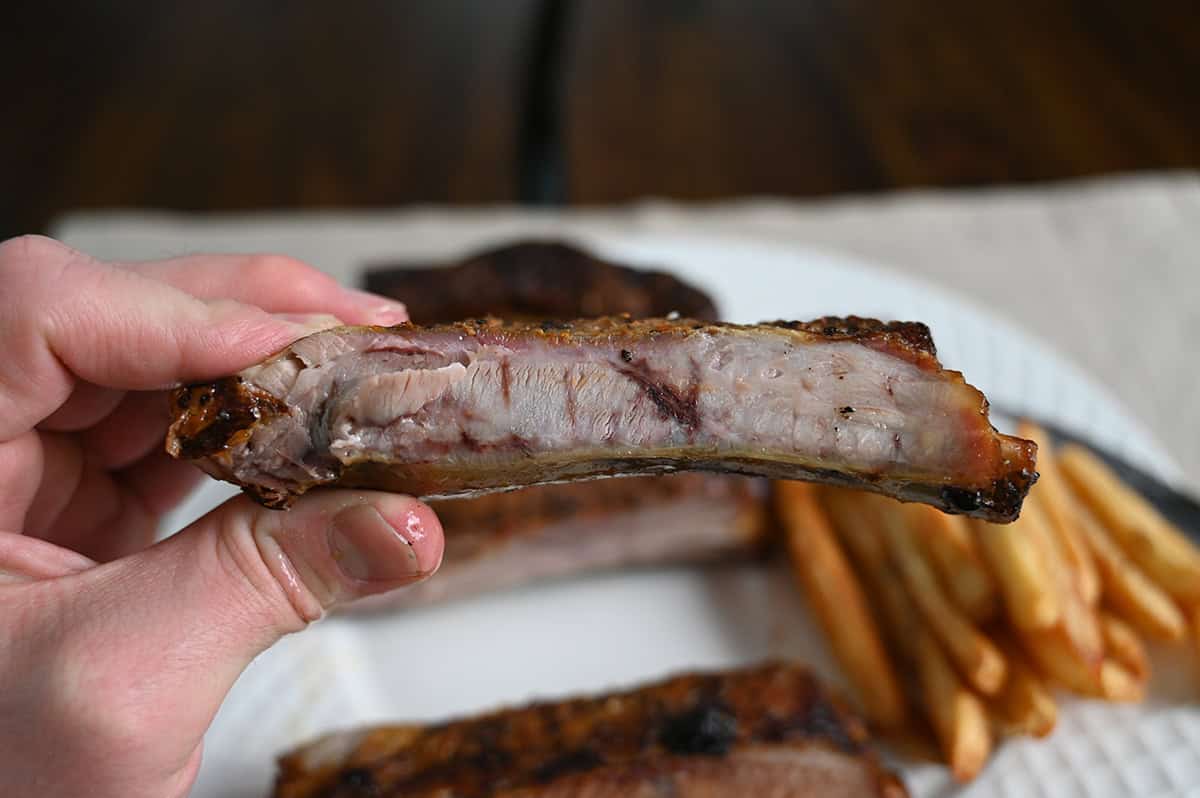 Closeup image of a hand holding one rib on it's side close to the camera so you can see the meat on the rib.