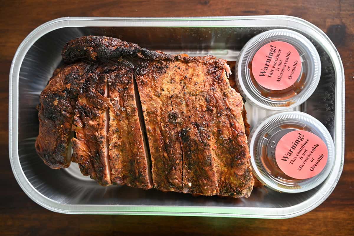 Top down image of an open tray ribs beside two small containers of barbecue sauce.