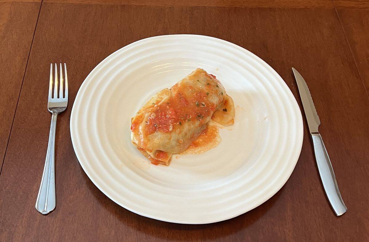 Image of one cooked cabbage roll served on a white plate with a fork and knife beside the plate.