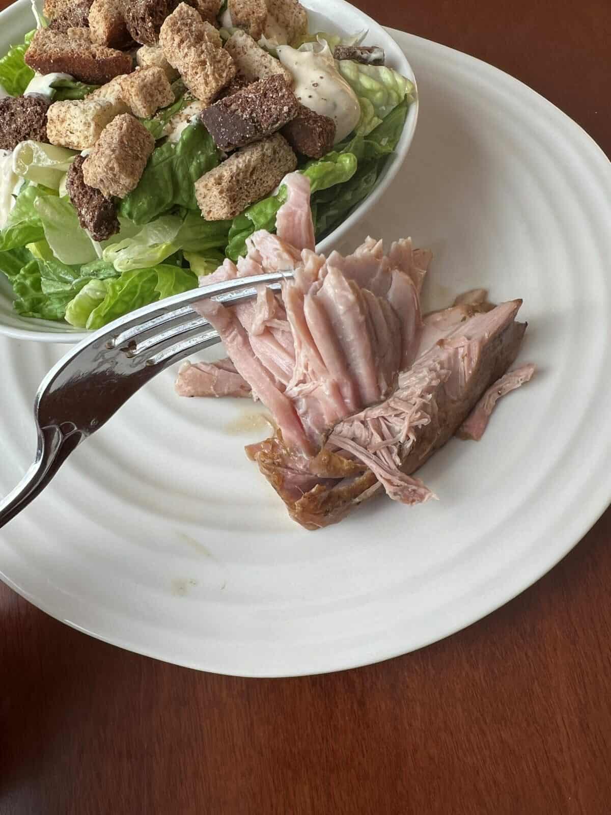 Top down image of a fork with pork on hovering over a plate with Caesar salad and pork roast.