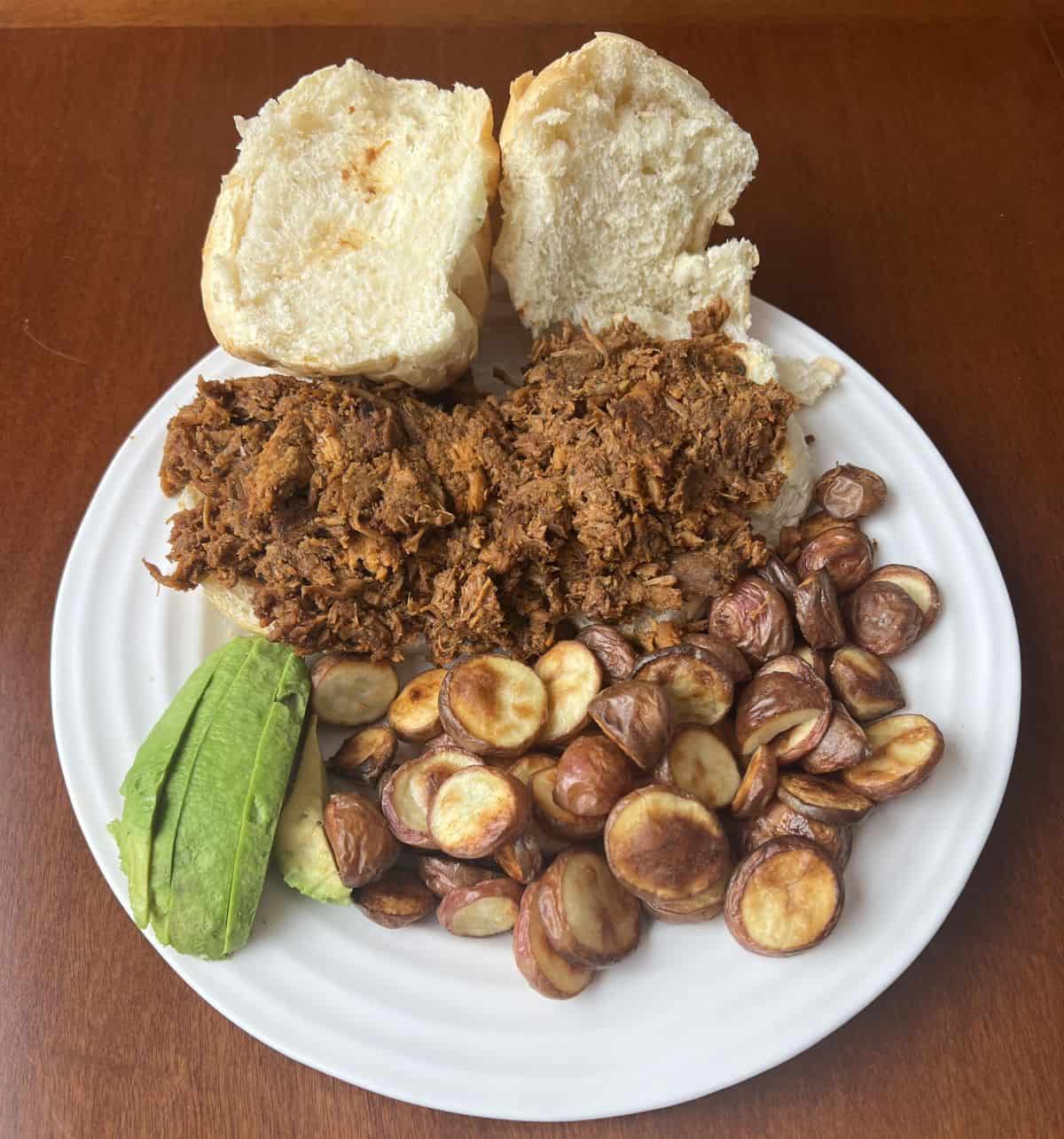 Image of the carnitas served on buns beside avocado and potato wedges on a white plate.