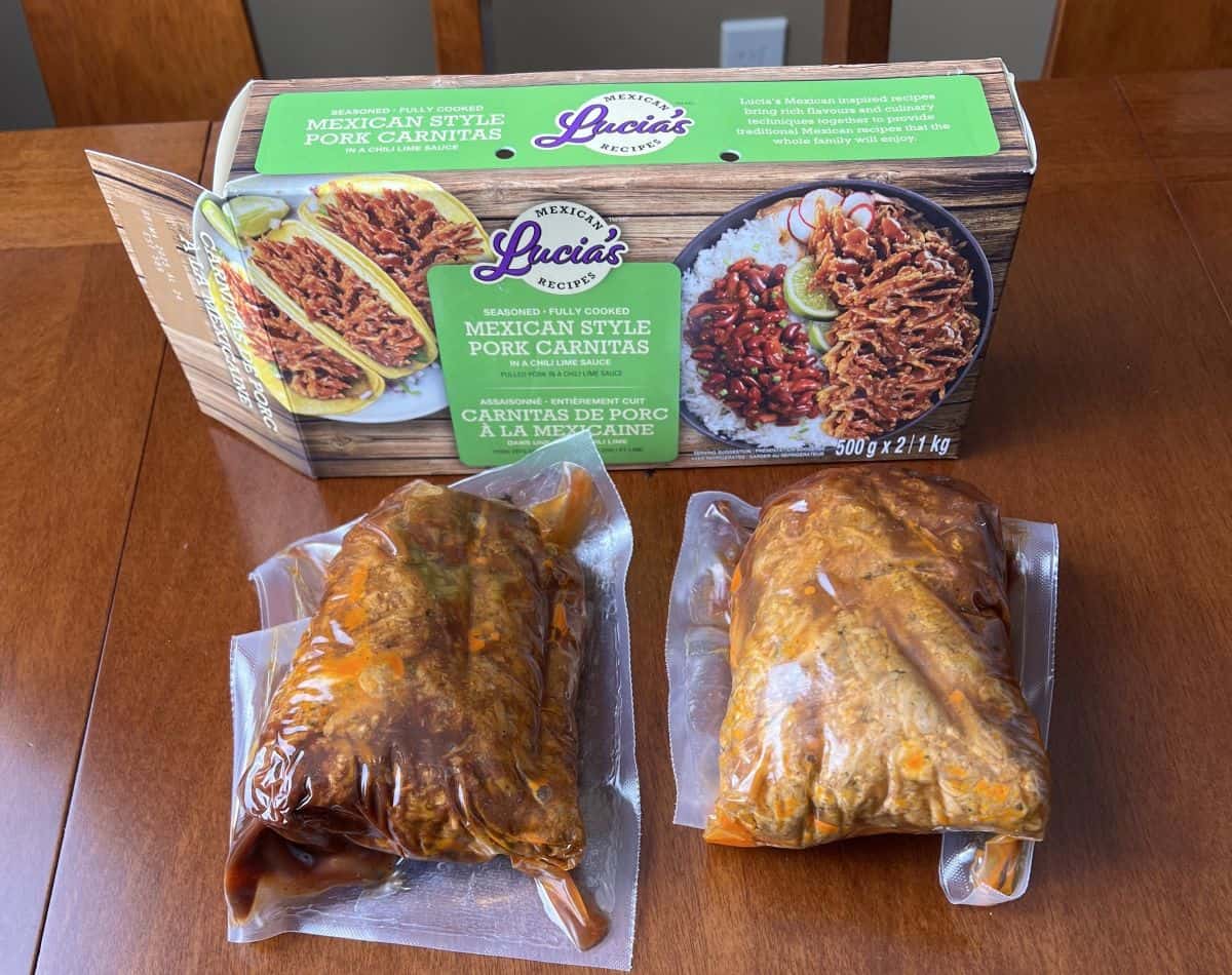 Image of two vacuum sealed packs of pork carnitas unopened in front of an open box of Lucia's Mexican Style Pork Carnitas. 