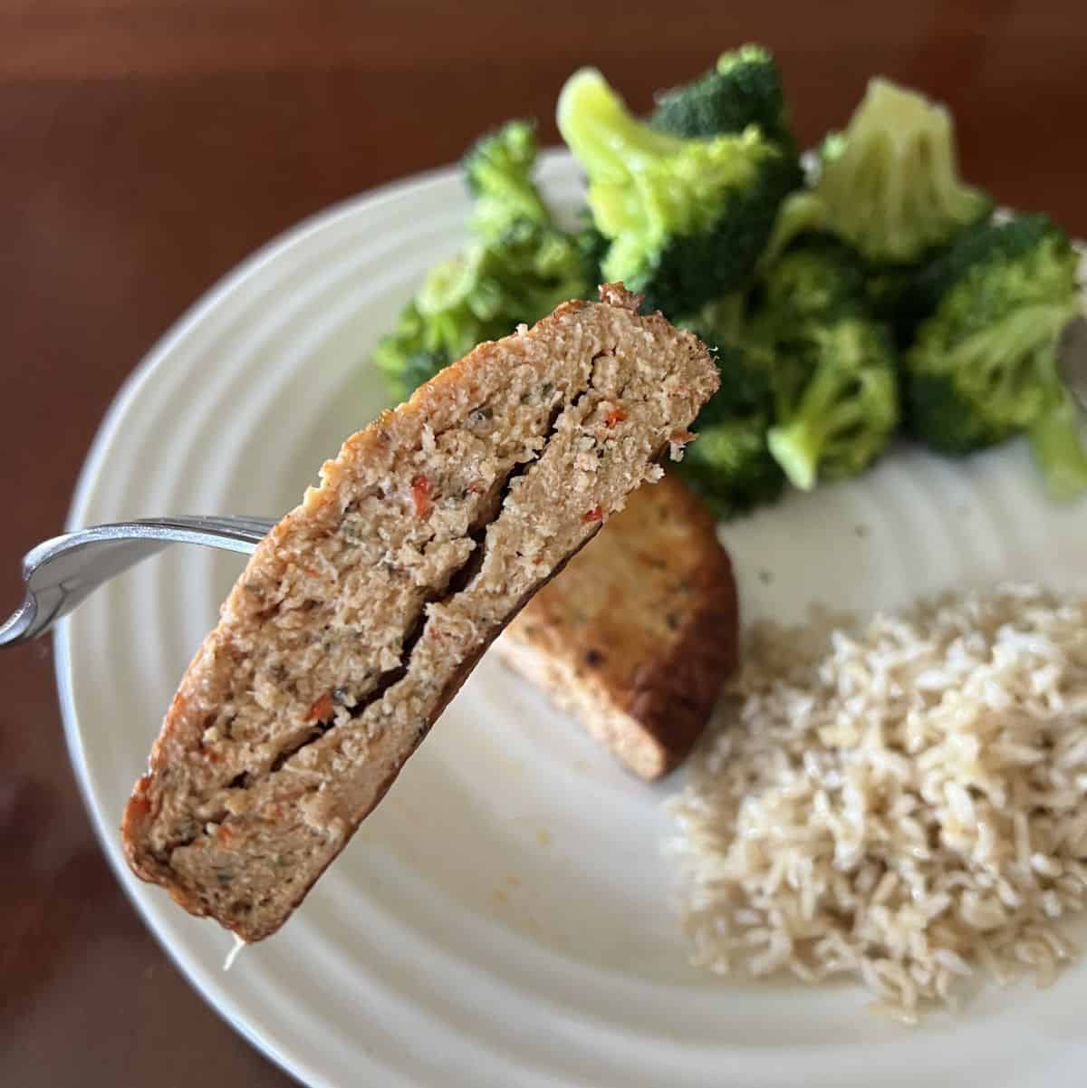 Image of a fork with a chicken burger cut in half on it hovering over a plate of rice and broccoli. 