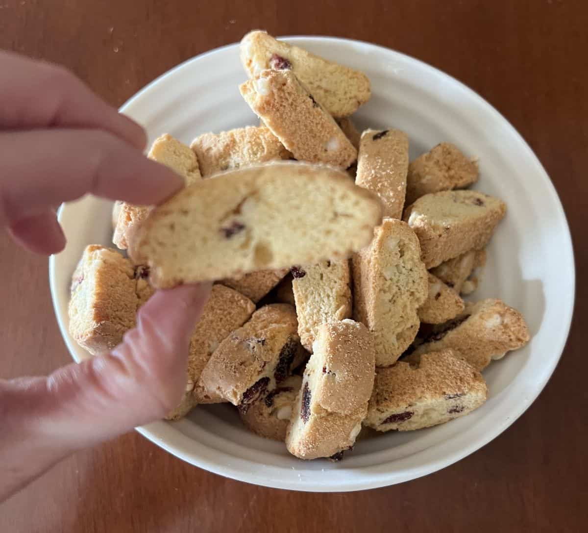 Image of a hand holding one cookie hovering over a bowl of Cooper Street Snacks Twice Baked Cookies. 
