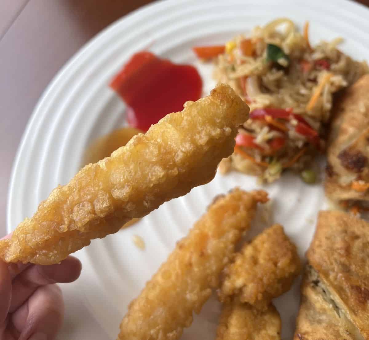 Image of a hand holding one tempura shrimp close to the camera with a plate of food in the background.