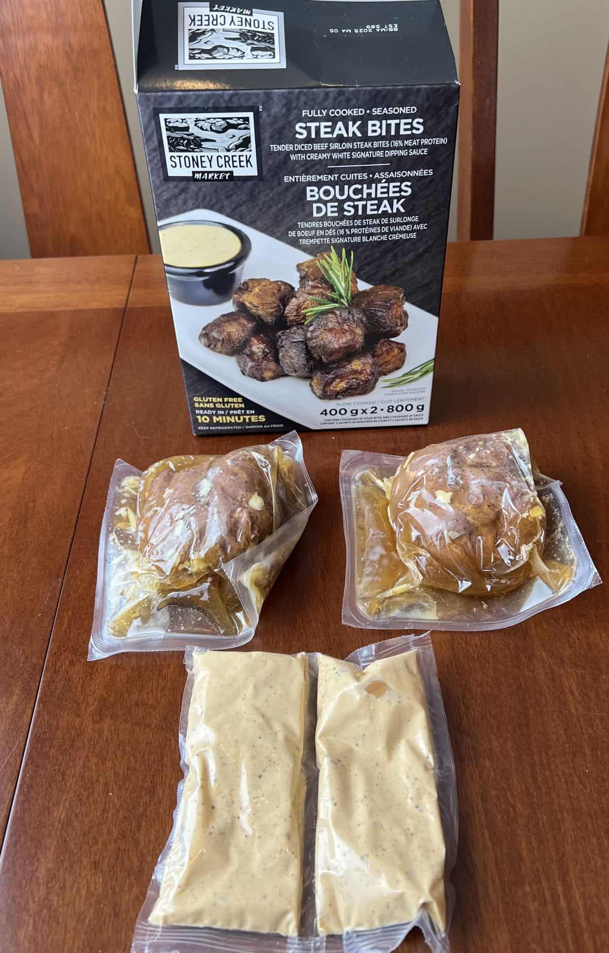 Image of the open box for the steak bites with two vacuum sealed packs of steak bites and two sauces in front of the box.