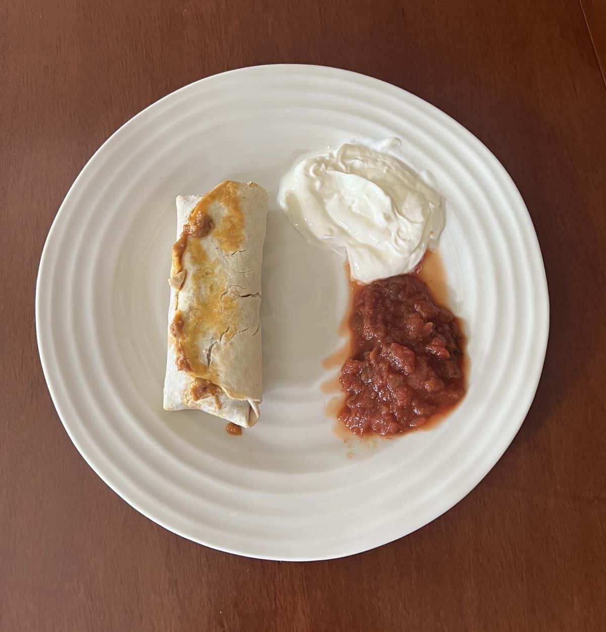 Image of one cooked burrito served on a white plate beside sour cream and salsa.