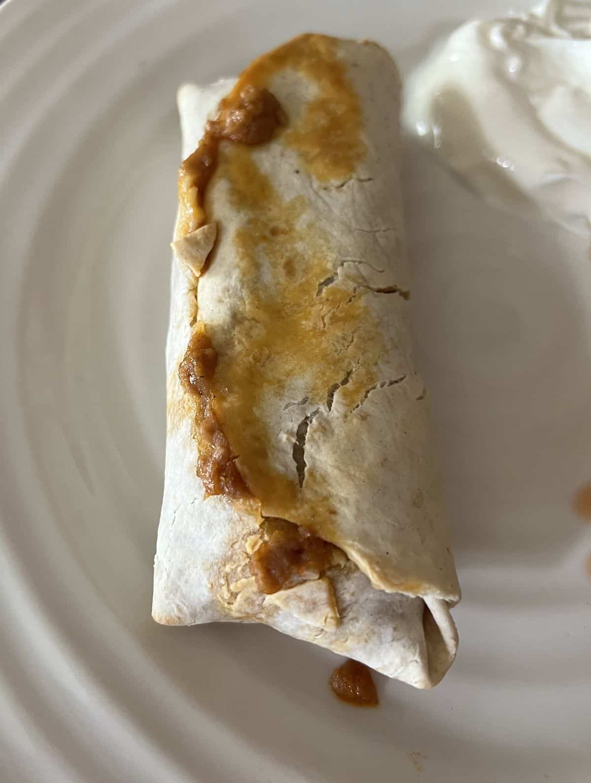 Image of one cooked burrito served on a white plate beside some sour cream. 