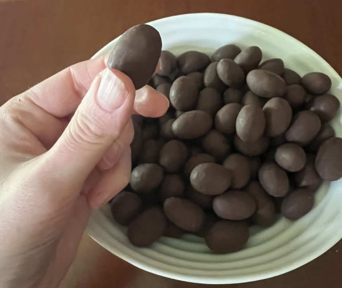 Image of a hand holding one Costco Edward Marc Chocolatier Coconut Almond above a bowl of coconut almonds.