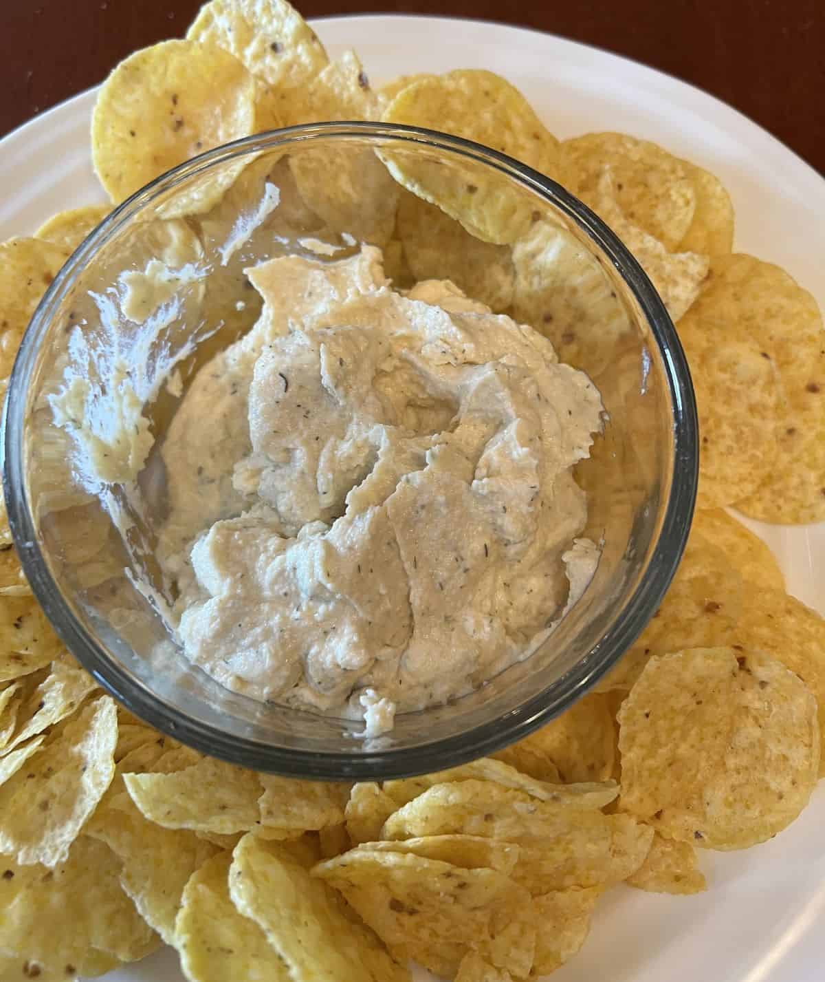 Top down image of a bowl of hummus in the middle of a plate with tortilla chips. 
