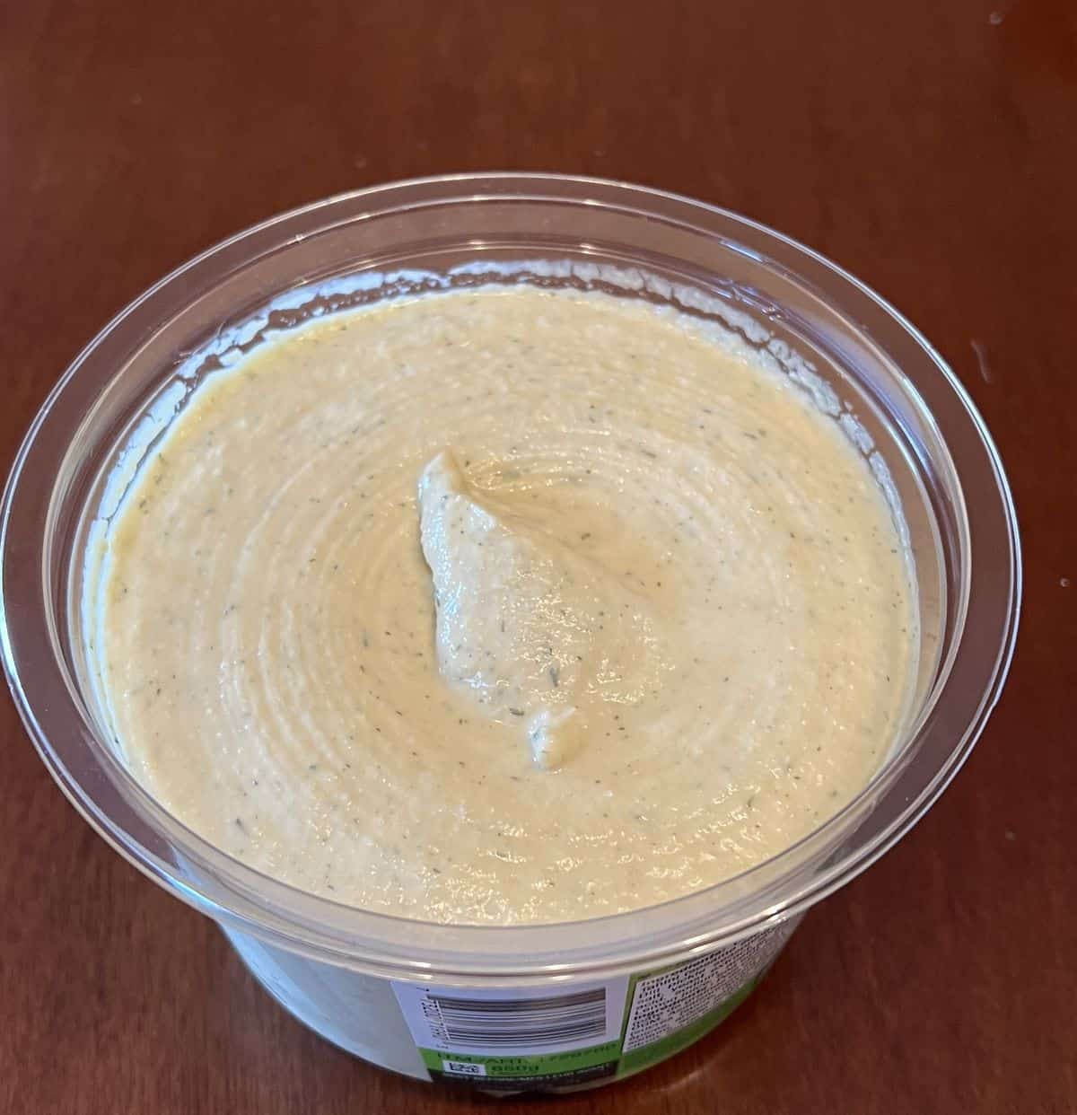 Image of a container of hummus with the lid off so you can see what the top of the hummus looks like. 