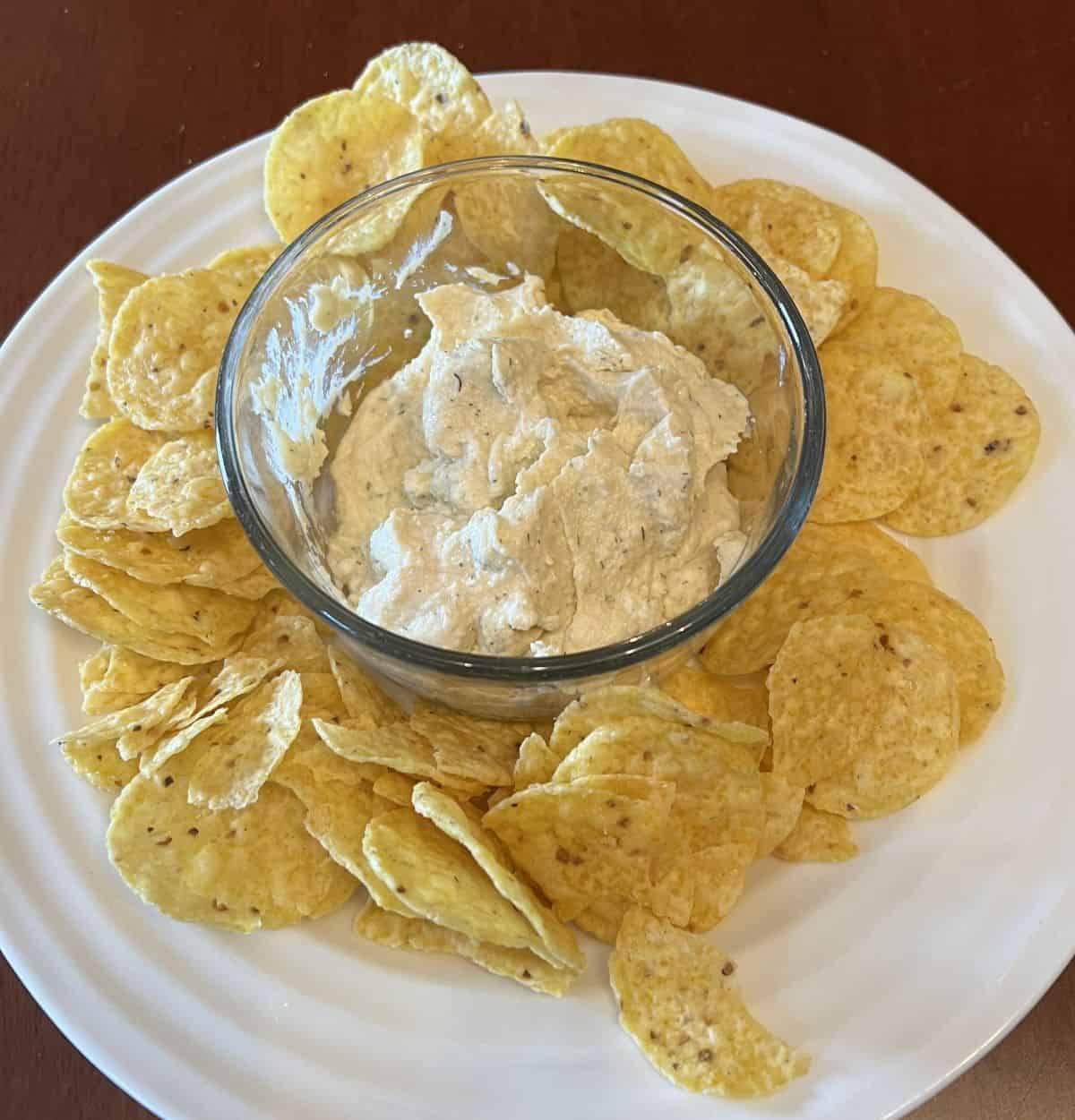 Image of a plate of tortilla chips with dill pickle hummus in a small bowl in the center of the plate. 