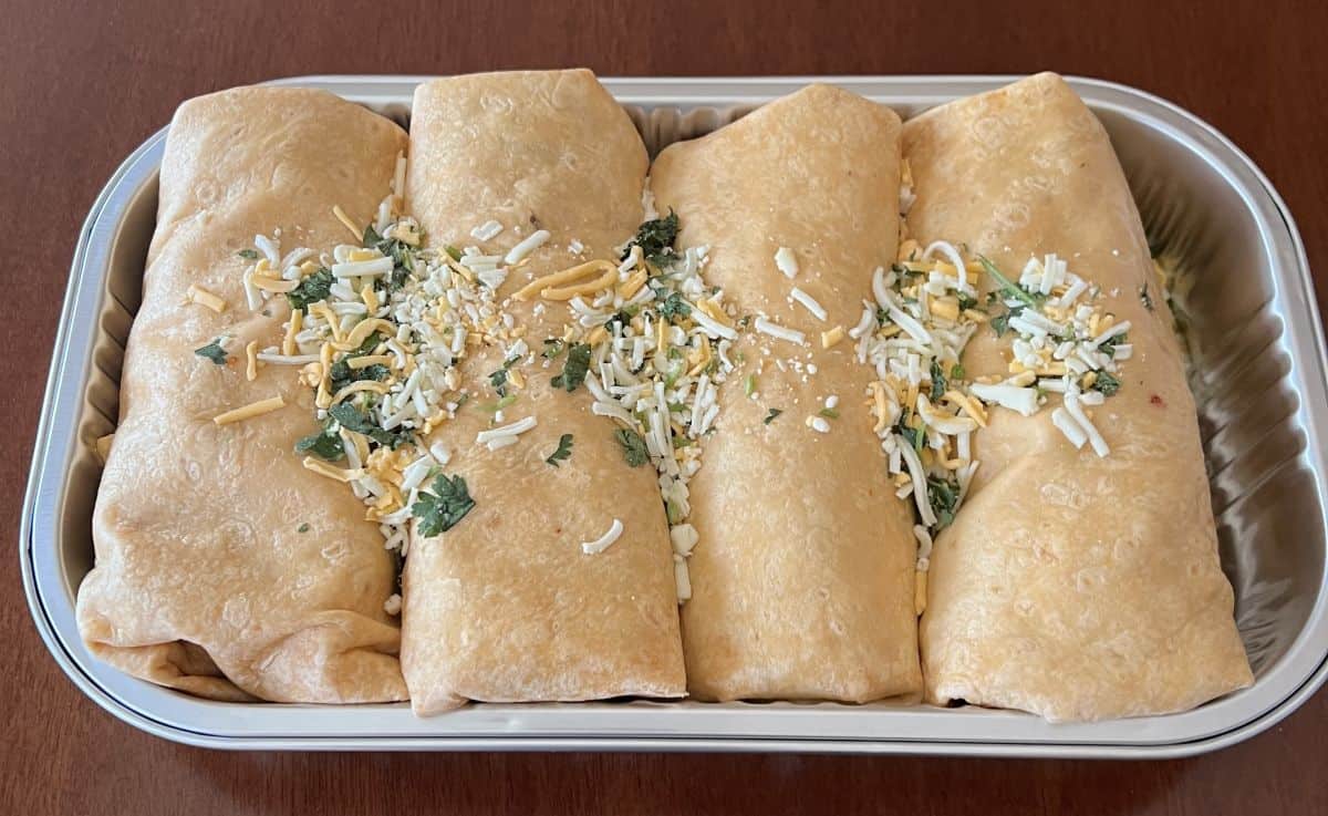 Image of four large burritos in a aluminum foil tray prior to heating them in the oven. There is cheese sprinkled on top. 