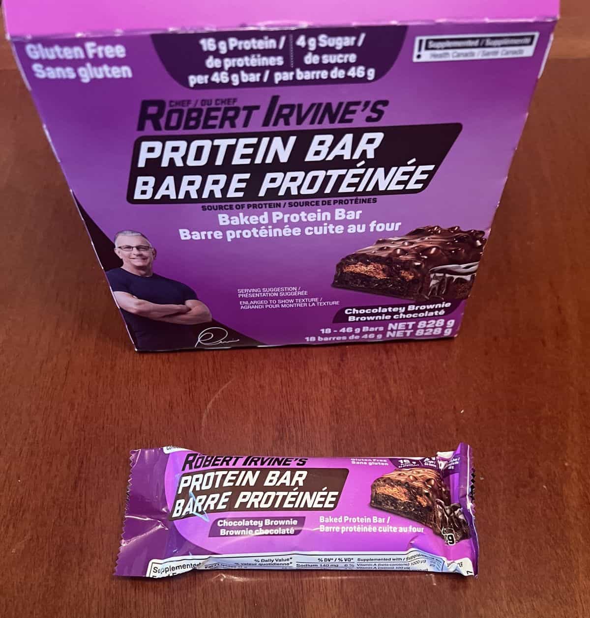 Image of one individually wrapped chocolatey brownie protein bar sitting on a table in front of the box of protein bars.