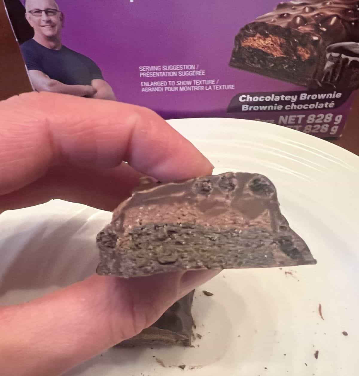 Closeup image of a hand holding one chocolatey brownie protein bar close to the camera and it's been cut in half so you can see the middle of the bar.