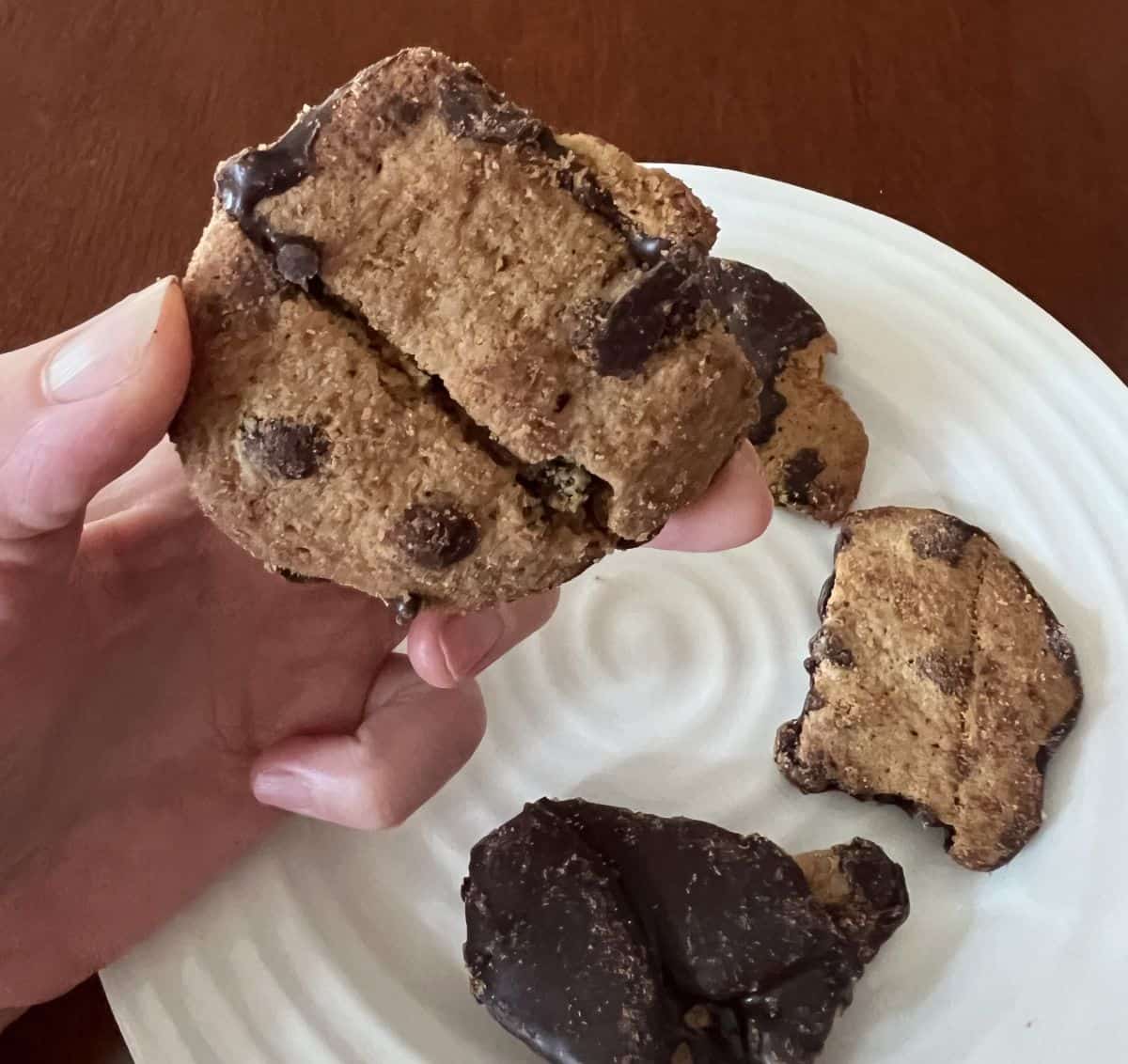 Image of a hand holding one piece of cookie bark close to the camera hovering over a plate of other cookie bark pieces.