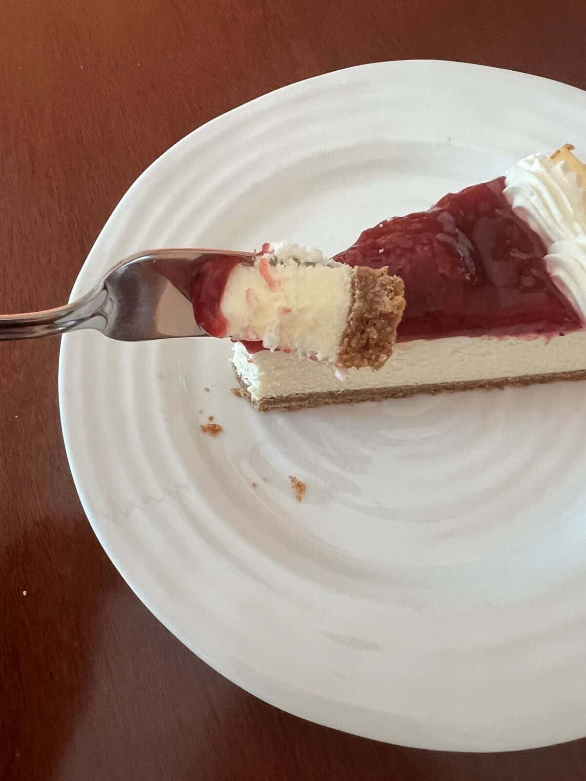 Image of a fork with a bite of cheesecake hovering over a plate with a slice of cheesecake on it. 