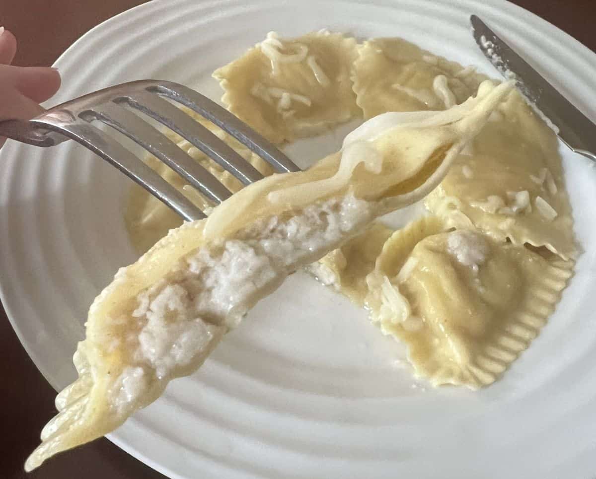 Image of a fork with a bite of ravioli on it hovering over a plate of ravioli. You can see the filling in the ravioli.