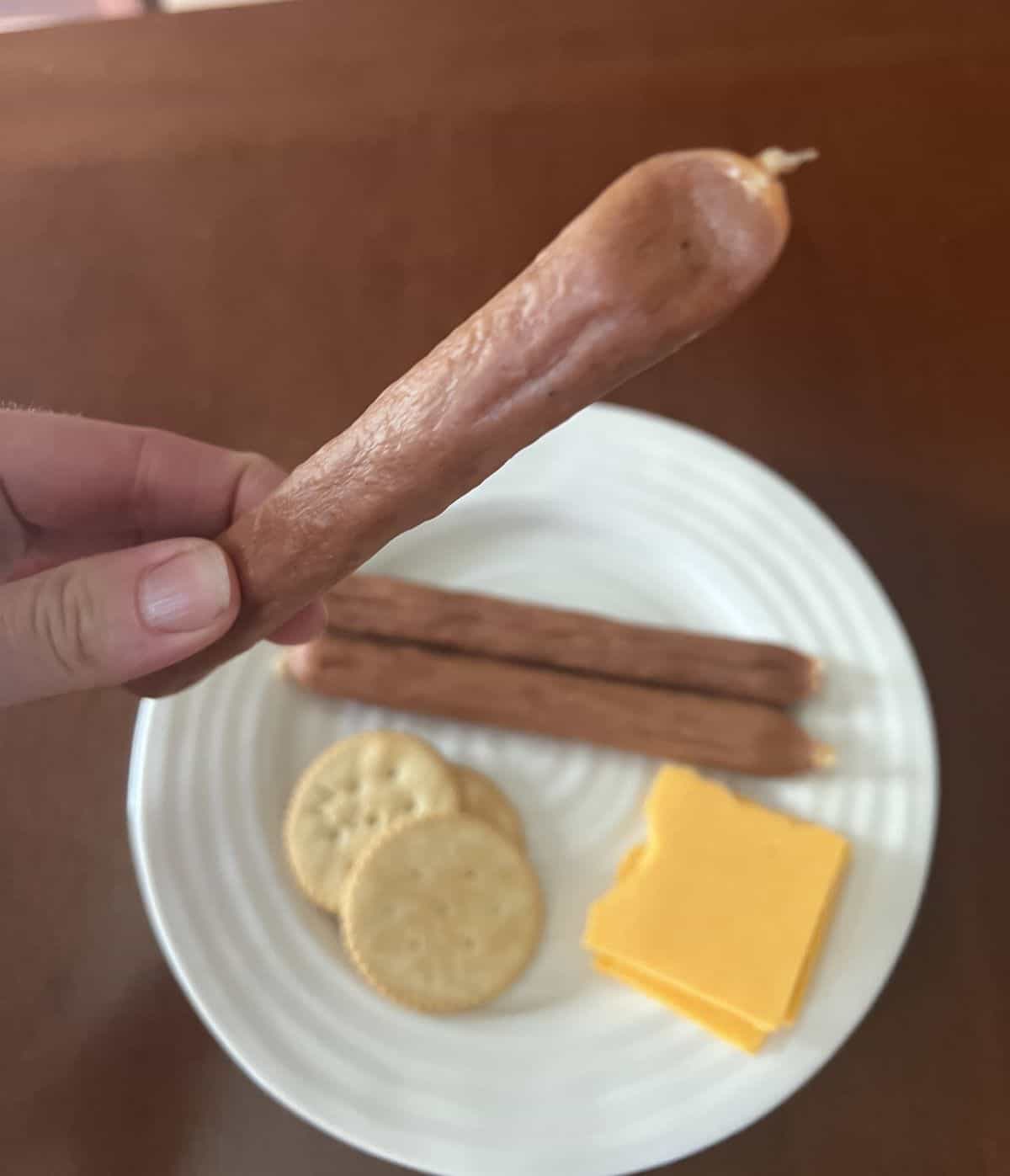 Image of three turkey pepperoni sticks served on a white plate beside cheese and crackers. A hand is holding up one turkey sausage. 
