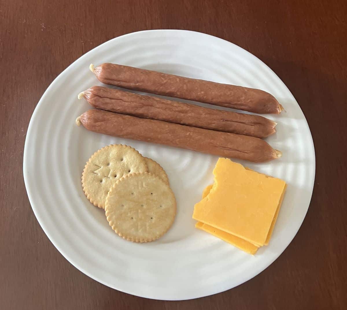 Image of three turkey pepperoni sausages served on a white plate beside cheese and crackers. 