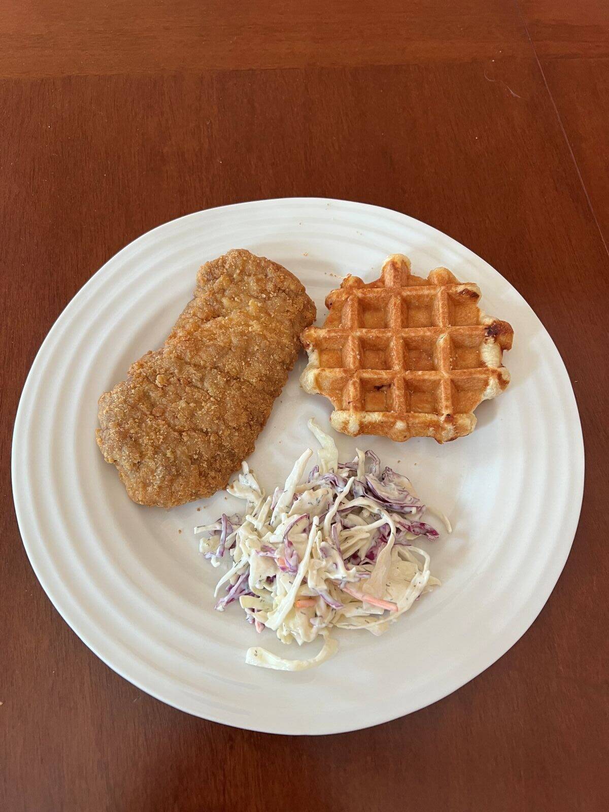 Top down image of a breaded chicken breast, waffle and coleslaw served on a white plate.