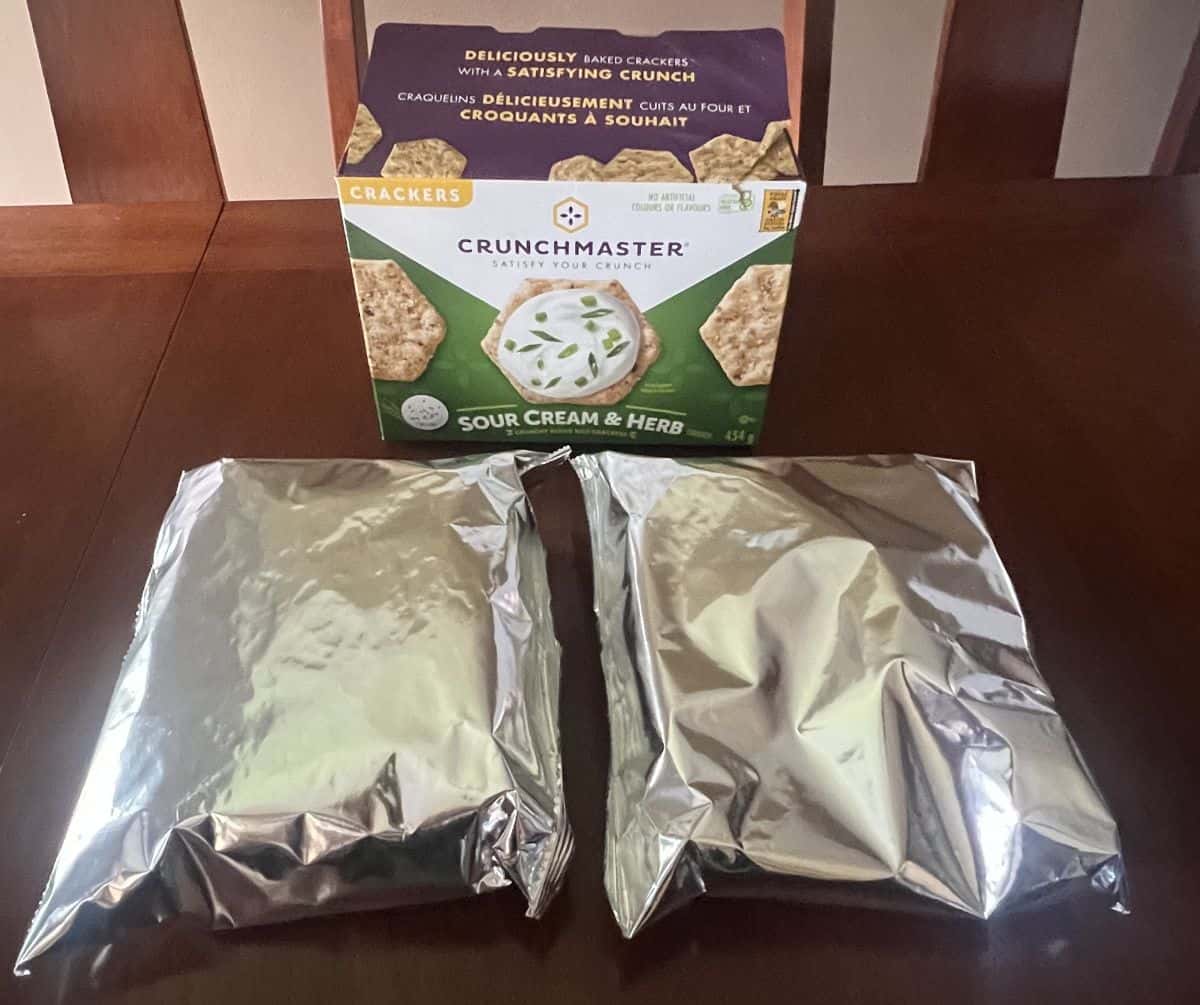 Image of two sealed bags of crackers in front of a box of the Costco Crunchmaster Sour Cream & Herb Crackers. 