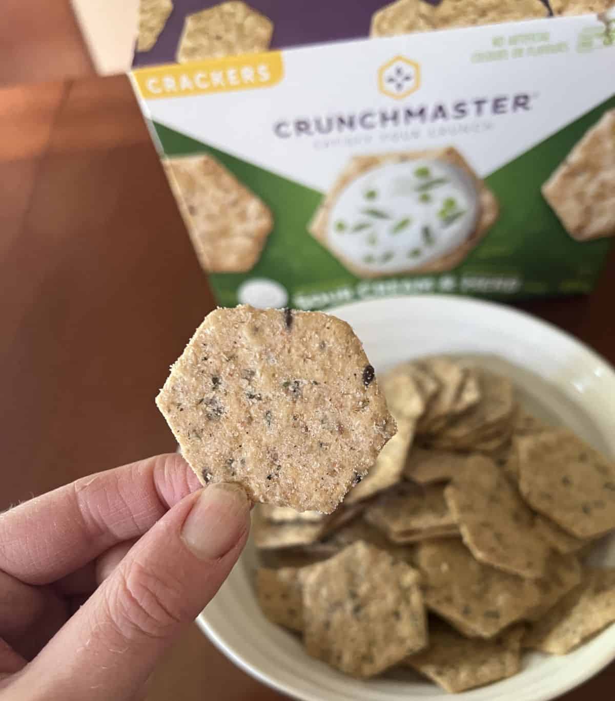 Image of a hand holding one sour cream & herb cracker hovering over a white bowl of crackers. 