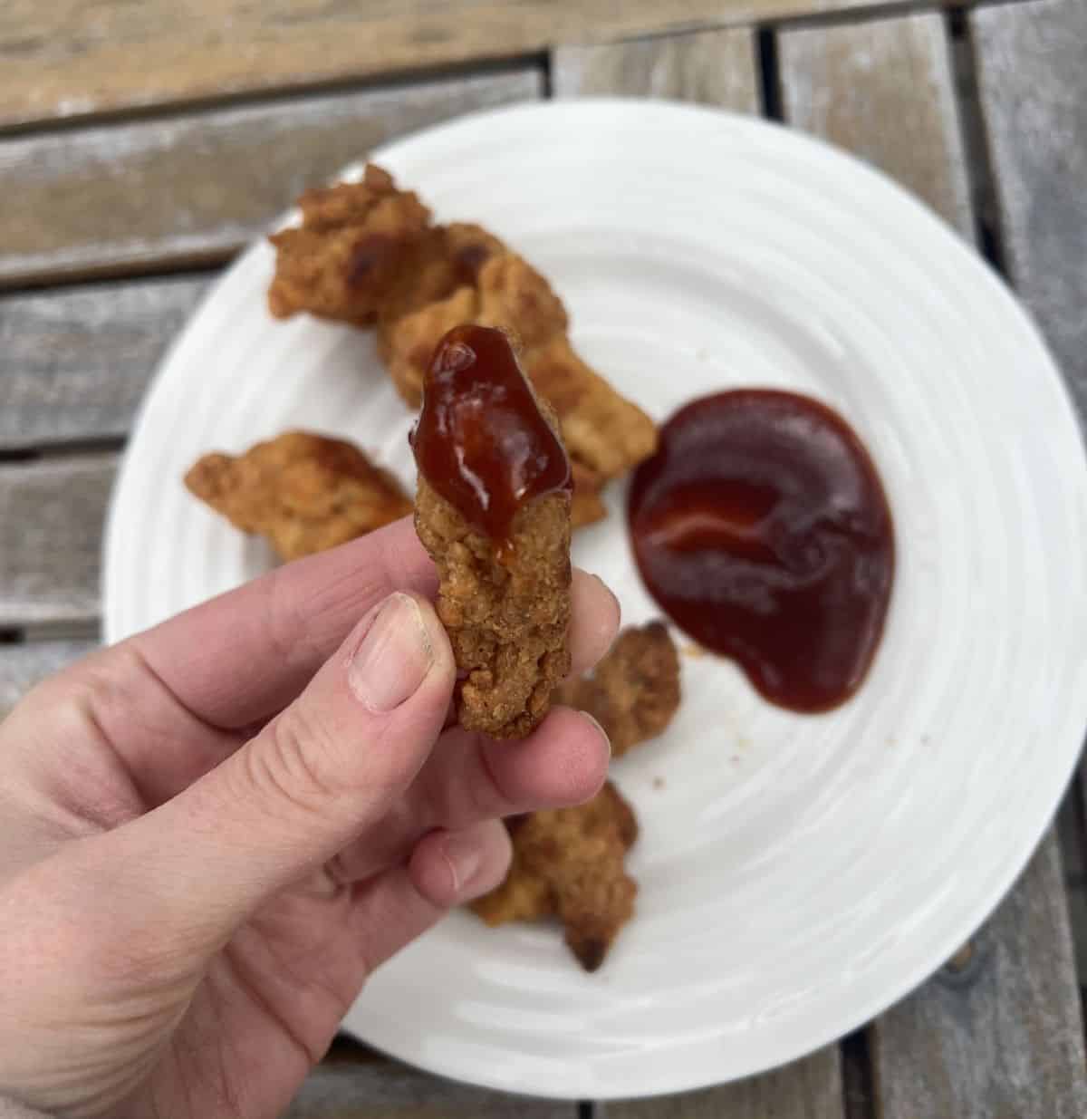 Image of a plate of chicken breast chunks with a side of barbecue sauce, there's a hand holding one chicken breast chunk dipped in barbecue sauce.