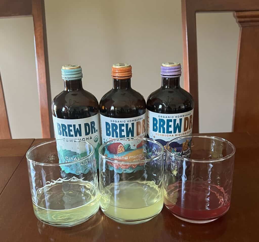 Image of three different kinds of kombucha poured into clear glasses sitting in front of the bottle of kombucha they came from. 
