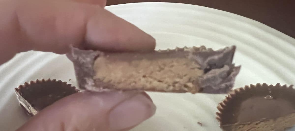 Closeup image of a hand holding one Reese's Peanut Buter Cup cut in half with caramel filling on the inside. 