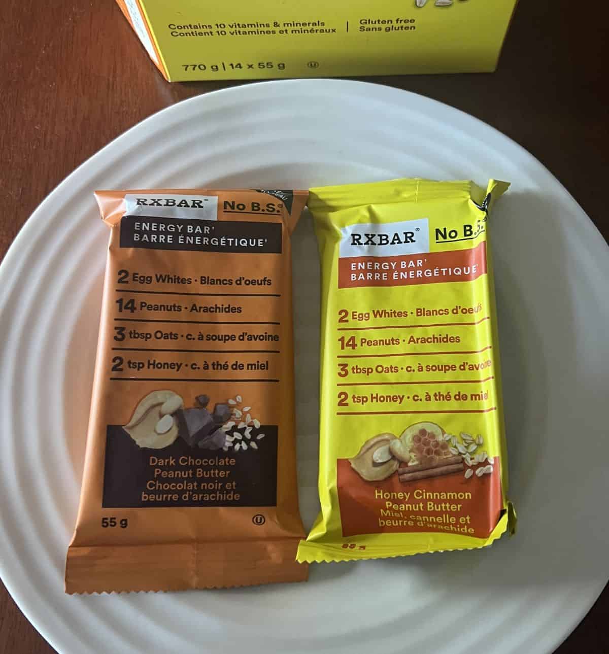 Image of one wrapped dark chocolate peanut butter bar beside one wrapped honey cinnamon peanut butter bar served on a white plate.