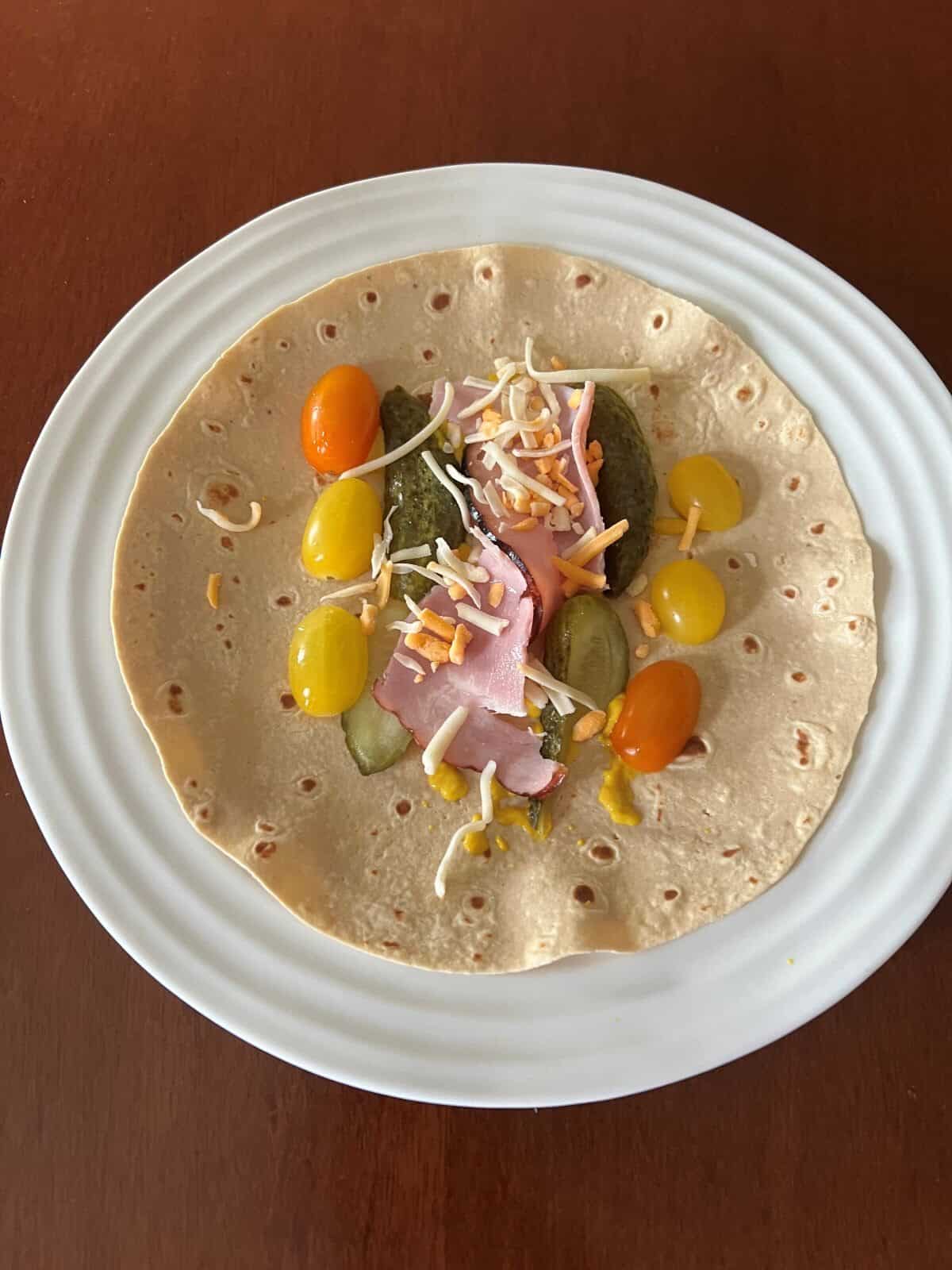 Image of one tortilla served on a white plate with toppings like ham, tomatoes, pickles on it.