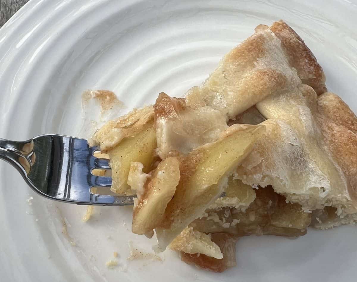 Image of a fork with a bite of apple pie on it hovering over a plate with a slice of apple pie on it. 