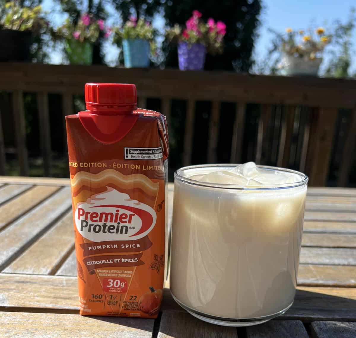Image of one pumpkin spice Premier Protein shake sitting beside a glass of protein shake with ice in it.