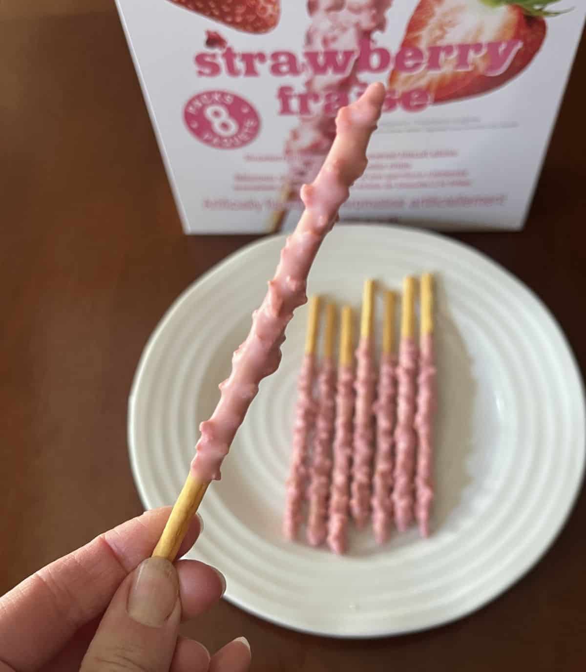 Image of a hand holding one Pepero biscuit stick close to the camera with a plate of Pepero in the background.