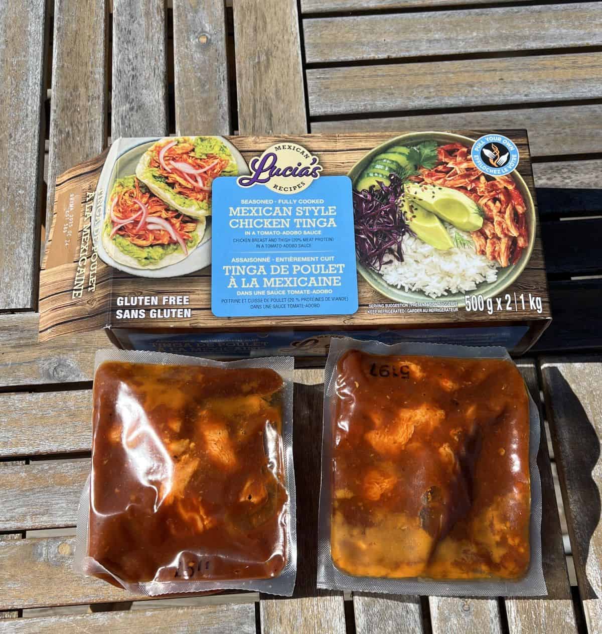Image of two vacuum sealed packs of chicken tinga sitting on a table in front of the box unopened. 