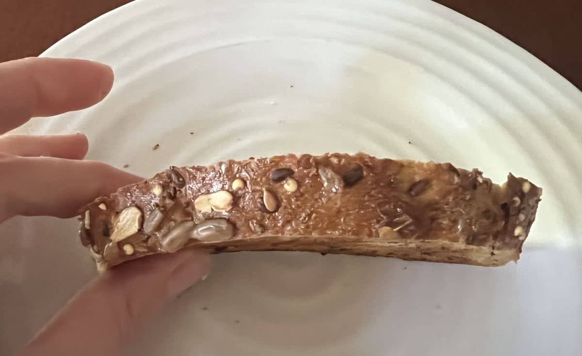 Image of a hand holding one slice of bread close to the camera on it's side so you can see oats and seeds on the crust of the bread.
